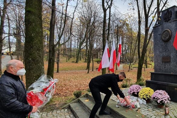 Dwie osoby, wójt i przewodniczący rady, skłądają kwiaty przed pomnikiem w parku. W tle drzewa