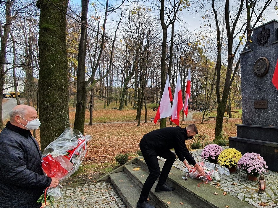 Dwie osoby, wójt i przewodniczący rady, skłądają kwiaty przed pomnikiem w parku. W tle drzewa