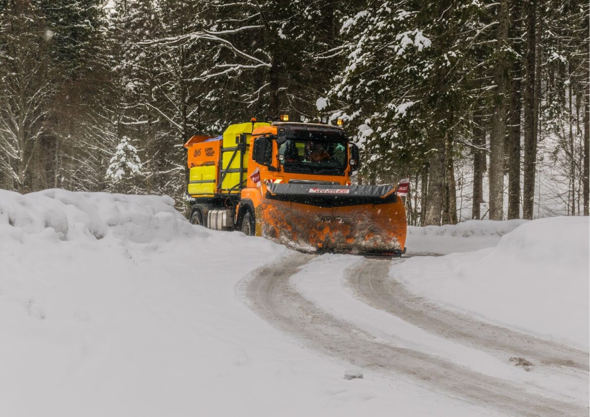Ciężarowy samochód z pługiem usuwa śnieg z drogi, W tle zaśnieżone drzewa