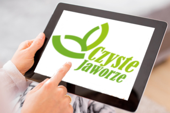 Ręka trzymająca tablet, drugą dłonią wskazany jest wyświetlacz z napisem "Czyste Jaworze"